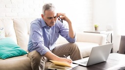 Man on phone in living room with laptop, pen, and paper
